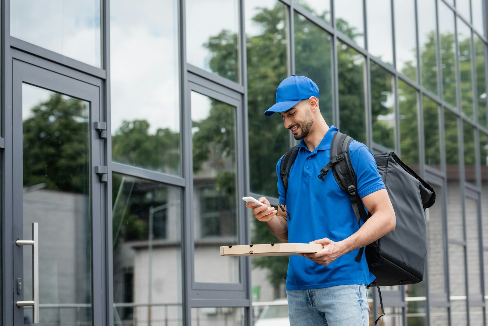 Smiling muslim courier with pizza box and smartphone standing near building outdoors